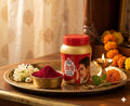 Jar of colored powder with a red lid on a tray with flowers and a candle, against a curtain background.