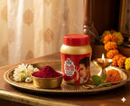 Jar of colored powder with a red lid on a tray with flowers and a candle, against a curtain background.