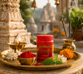 Red container labeled 'Bhagvati' on a tray with flowers, incense, and temple in the background